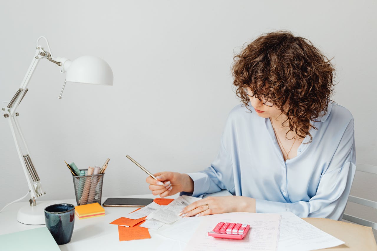 Woman in office calculating expenses with documents, calculator, and coffee.